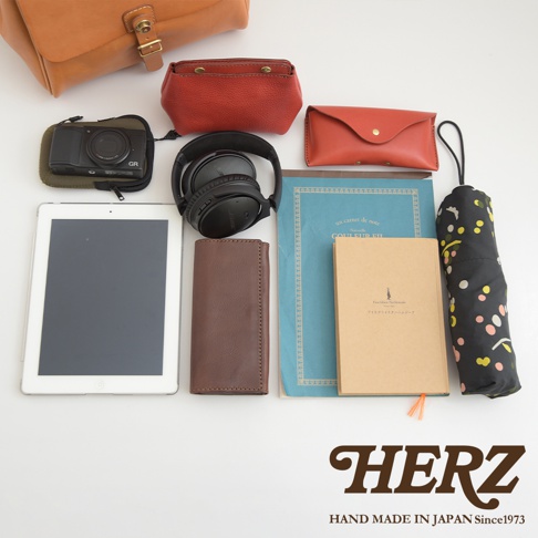 職人手作皮革屋瓦風後背包 R-15，展示內容包含：棕色皮革翻蓋後背包、紅色皮革化妝包、紅色皮革證件夾、黑色數位相機、黑色頭戴式耳機、白色平板電腦、棕色皮革長夾、藍色筆記本、米色筆記本，以及印有彩色圓點圖案的黑色摺疊傘。