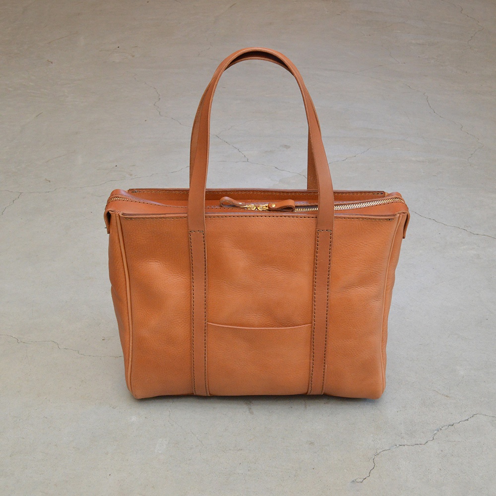 一只棕褐色皮革製成的托特包，配有兩個提把和一個拉鍊封口。包包正面有一個貼袋，整體呈現簡約風格。