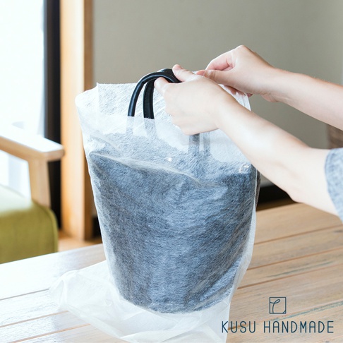 一雙手正在將一個黑色、材質類似毛氈的圓筒形物品放入透明的防塵套中，防塵套上方有兩個黑色提把，物品和防塵套皆置於木製桌面上。