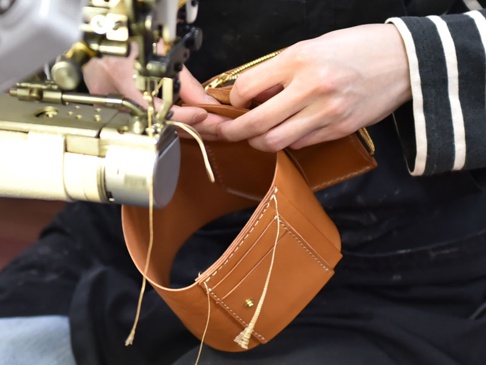 職人正在使用縫紉機製作一個棕色皮革製的直式卡片收納雙折短夾，皮革表面有縫線和卡槽細節。