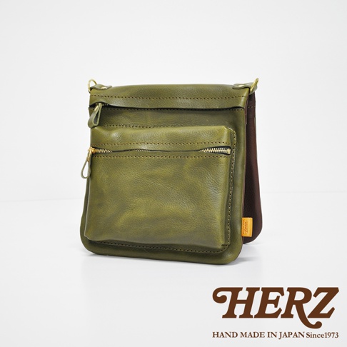 職人手作皮革翻轉雙色直式斜背包，橄欖綠色，具有金色拉鍊和金屬扣環，皮革材質，背面有品牌標籤。