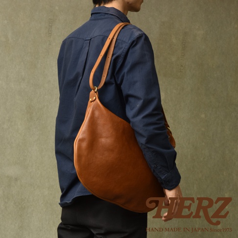 一個男性模特斜背著棕色皮革製的半月形單肩包，包包呈現自然的皺褶，搭配深藍色襯衫與黑色褲子。