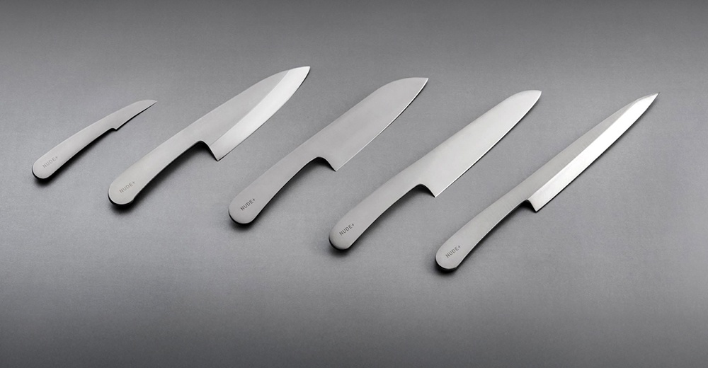 志津匠 Nude+ 系列共有五把金屬質感的廚房刀具，從左至右依序為削皮刀、主廚刀、日式廚師刀、三德刀與切片刀，擺放於灰色背景上。