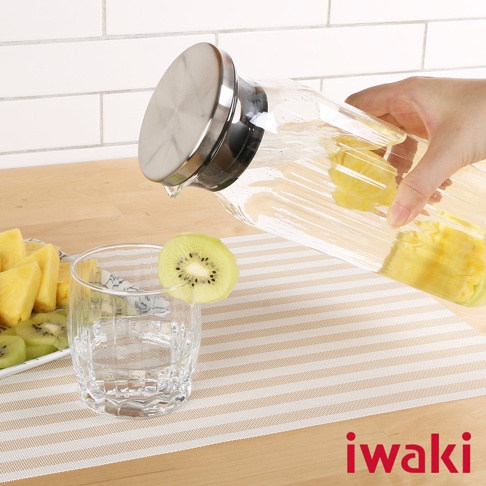 透明玻璃水壺，帶有不鏽鋼蓋，正傾倒出含有鳳梨塊的飲品。一旁有切片奇異果裝飾的玻璃杯，以及盛有鳳梨塊和奇異果片的盤子。