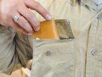 淺棕色皮革箱型零錢包，邊緣有白色縫線，置於卡其色襯衫的胸前口袋中，一只戴著銀色笑臉戒指的手正在將零錢包放入口袋。