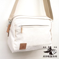 一個米白色帆布斜背包，正面有拉鍊口袋，右下角有一個棕色皮革標籤，上面有圖案和文字，旁邊還有東京淺草犬印鞄製作所的圖案和文字。