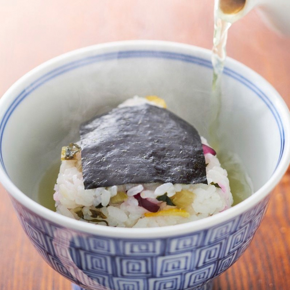 一個裝有白米飯、海苔和醃漬配料的碗，上方倒入熱湯。碗內有藍白色的幾何圖案，碗邊緣有兩條藍色細線。