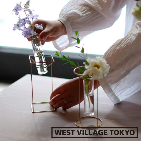 黃銅幾何框架支撐玻璃試管花器，其中一株插入紫色小花，另一株則插有白色花朵與綠葉。