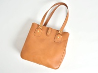 淺棕色皮革手提包，有兩個短提把，包身中央有搭扣設計，邊緣以白色縫線裝飾，風格簡約。