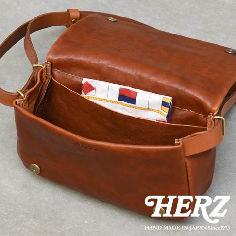 職人手作皮革斜背包，呈現自然的棕褐色，包身線條簡潔，翻蓋設計，內層可見印有航海信號旗圖案的布料，金屬釦件點綴。