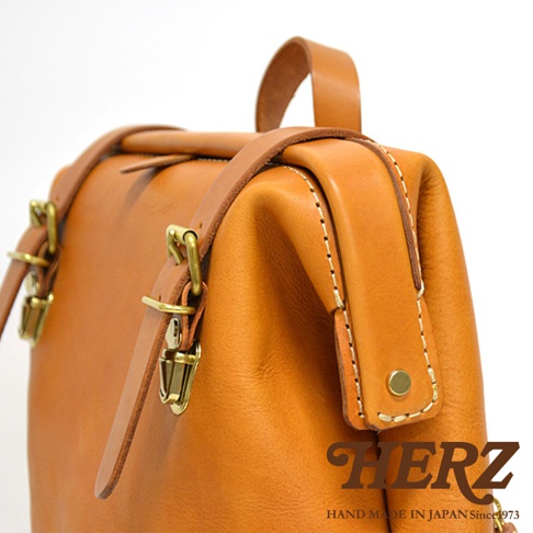 職人手作皮革後背包，採用溫暖的橘棕色皮革，搭配復古金色金屬扣環與皮帶，展現精緻車縫工藝。
