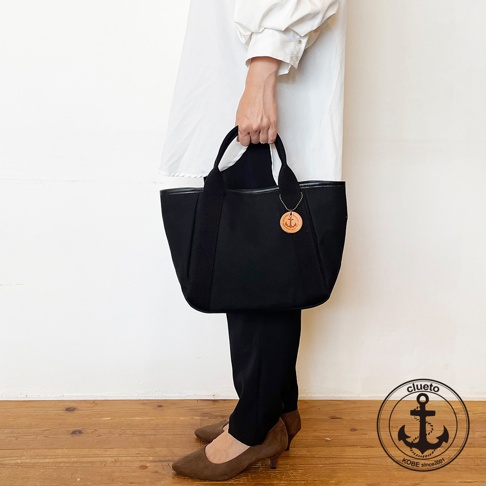 黑色帆布手提托特包，配有黑色織帶提把，包身側面有黑色皮革裝飾，包上掛有圓形木質吊飾，上面刻有船錨圖案。
