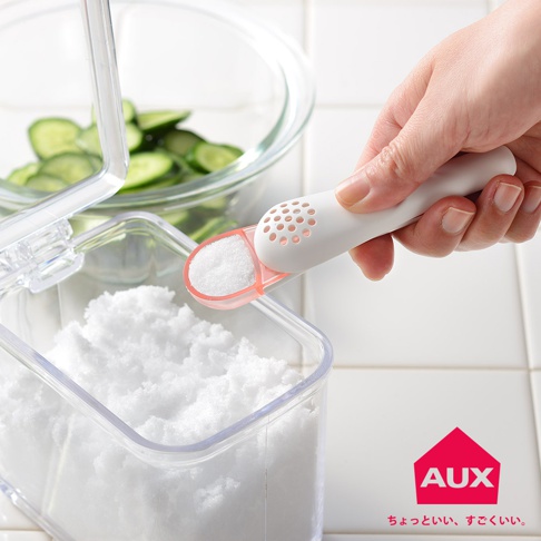 白色帶有圓孔的鹽匙，匙頭為粉紅色，正從裝有大量白色細鹽的透明容器中舀取鹽。背景有裝有切片小黃瓜的玻璃碗。