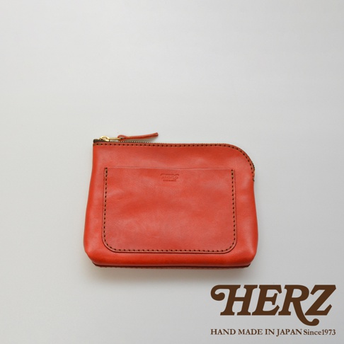 橘紅色皮革小物收納包，正面有一個貼袋，配有金色拉鍊，邊緣以深色縫線勾勒，展現皮革紋理。
