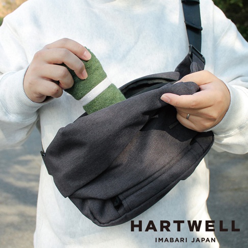 一個人將一個捲起來的綠色毛巾放入一個深灰色的斜挎包中。毛巾上綁著一條白色的束帶，上面印有 soto 字樣。包包的底部有 HARTWELL IMABARI JAPAN 的字樣。