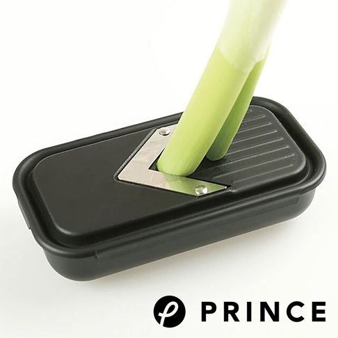 黑色塑膠製的磨泥切片兩用盒，內含不鏽鋼磨片，一根綠色蔥段正在通過磨片。