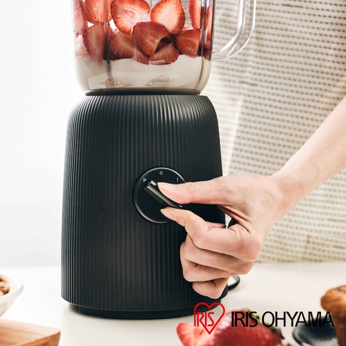 黑色直條紋紋理的果汁機/料理機，搭載透明果汁壺，壺內有切片草莓與奶昔。一隻手正在操作機身上的黑色旋鈕，旋鈕上有「P」、「O」、「I」等標示。