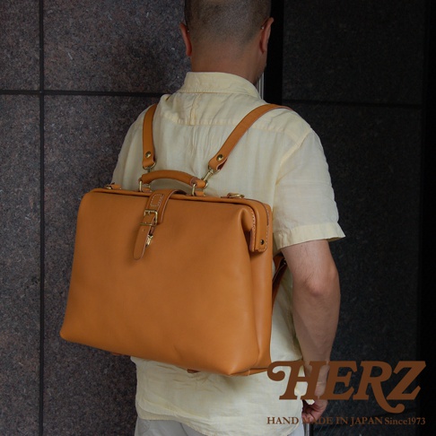 職人手作皮革軟式杜勒斯包，採用棕褐色牛皮製作，包包設計簡約，配有頂部提把、可調節肩帶及金屬扣環。