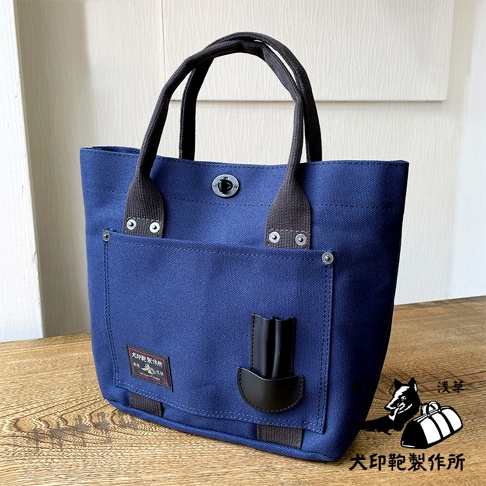 藍色帆布水桶托特包，配有深灰色提把，正面有一個大口袋，口袋上附有黑色皮革裝飾和一個紅色標籤。