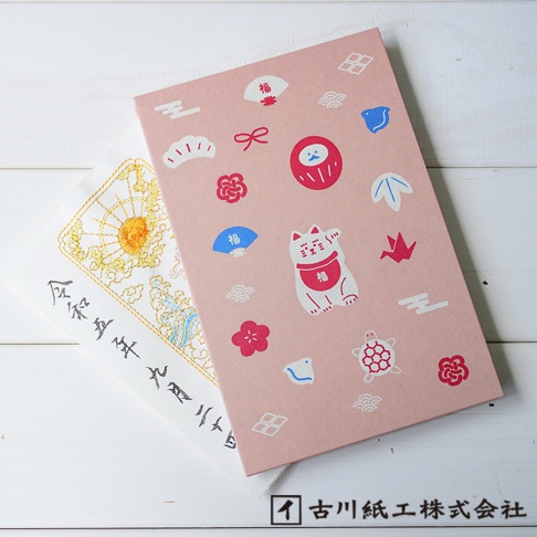 粉色和紙封面印有白色、紅色、藍色的吉祥物圖案，包含達摩、招財貓、烏龜、扇子、花朵、雲朵、紙鶴及日式幾何圖案。左側疊放著一本白色繡花御朱印帳，繡有金色太陽和祥雲圖案，並有日文書法。
