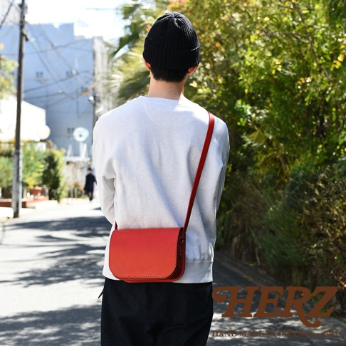 職人手作紅色皮革翻蓋橫式單肩包，有著簡約的設計和精緻的縫線。包款由高品質皮革製成，配備一條細長的紅色肩帶，可斜挎於身體一側。