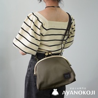 從背面拍攝，一位女子穿著條紋上衣和黑色牛仔褲，斜背著一個軍綠色的大玉口金斜背包，包上有 AYANOKOJI 的品牌標籤。