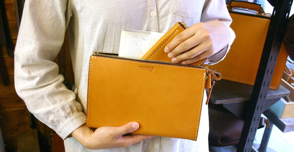 職人手作皮革三角底拉鍊手拿包，淺棕色皮革，搭配米色縫線，拉鍊開口，包身正面有「HERZ」壓印。