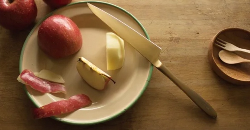 淺金色金屬水果刀，刀刃鋒利，刀柄為淺棕色，放置於裝有蘋果塊、蘋果皮和兩個紅蘋果的盤子上。右側有一個木碗，內有木製叉子和湯匙。