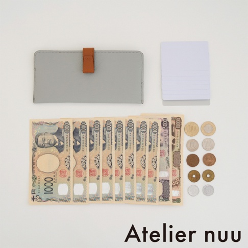 一個灰色皮革製成的 Atelier nuu 長夾平放於白色背景，附帶一個棕色皮質扣環。長夾上方整齊疊放著五張白色空白卡片，下方則排列著八張面額為一千日圓的紙鈔、一張五千日圓紙鈔與一張一萬日圓紙鈔，右側還排列著十枚不同面額的日本硬幣，整體呈現簡潔俐落的收納展示。