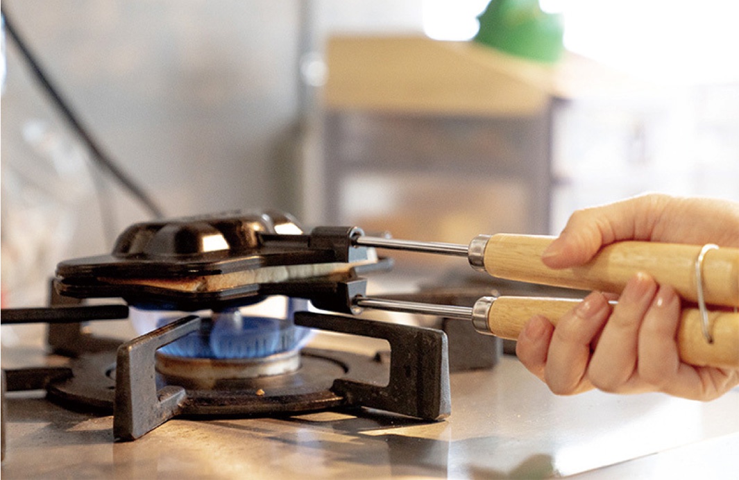 手持木柄熱壓三明治烤夾，正在瓦斯爐上烘烤三明治。烤夾為黑色，帶有銀色金屬連接處和圓形木製握把。