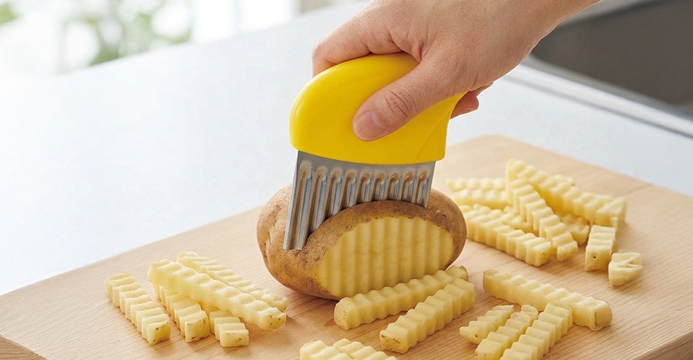 一隻手握著黃色握柄的下村工業波浪造型切刀，正在砧板上將馬鈴薯切成波浪狀的長條，一旁已擺放許多切好的薯條，呈現專業料理工具的使用情境。
