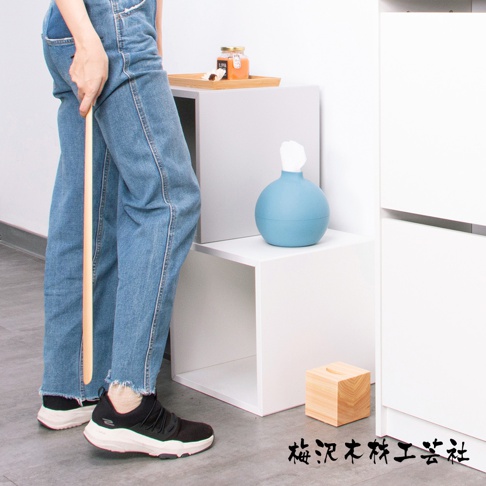 加長檜木鞋拔，末端有一個弧形握柄，整體為原木色。旁邊擺放著一個藍色圓形衛生紙盒和一個木製面紙盒。