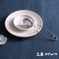 銀色不鏽鋼製的泡茶器，帶有網狀濾網和長柄夾子，放置於白色盤子上，背景為藍色紋理布墊。