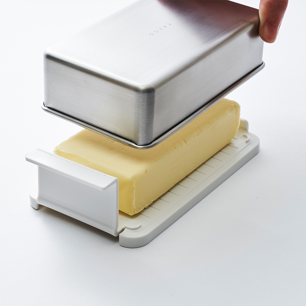 日製奶油盒，包含一個長方形的黃色奶油塊，放置在一個淺灰色、帶有紋路的底座上。銀色金屬製的盒蓋，印有「FAICO」字樣，正被一隻手拿起，準備蓋上。