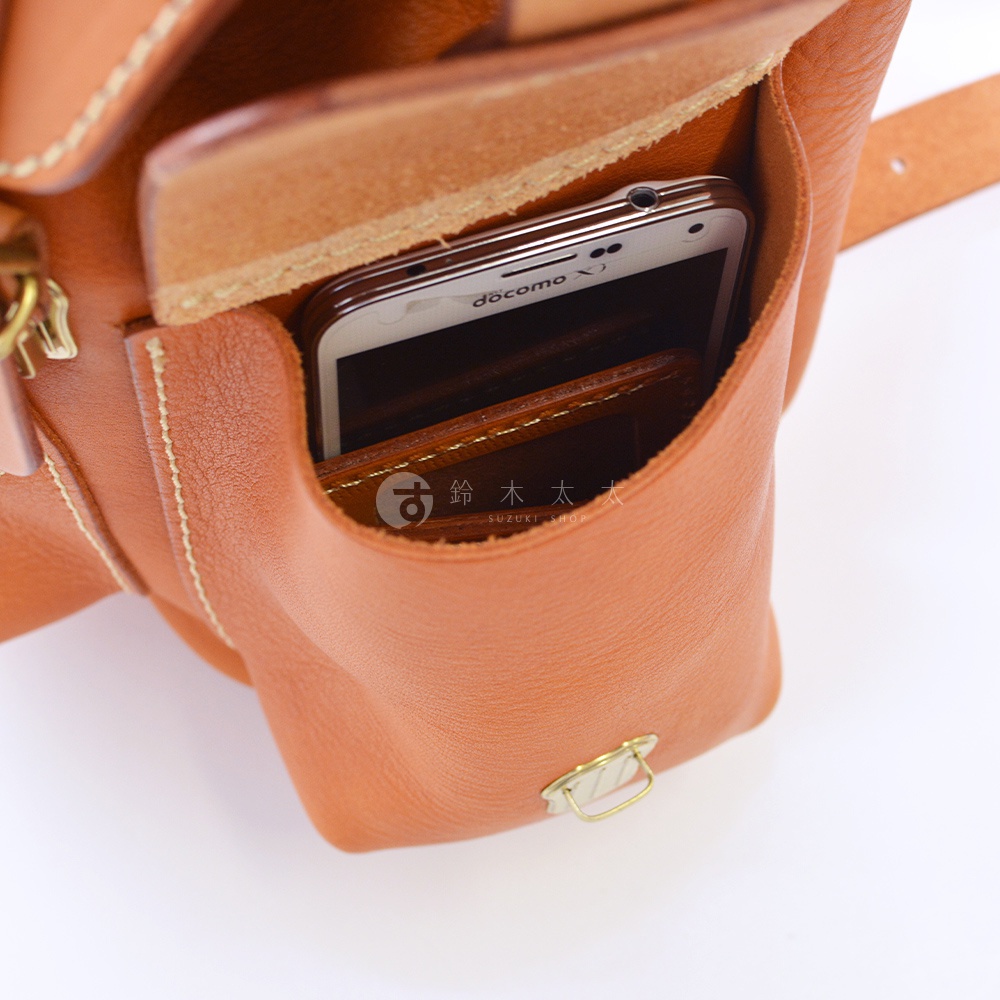 職人手作皮革復古風旅行後背包 R-65，特寫鏡頭顯示背包側邊的開放式口袋，一個白色手機和深棕色皮夾被放入其中，背包採用棕褐色皮革，搭配米白色縫線和金色金屬扣環。