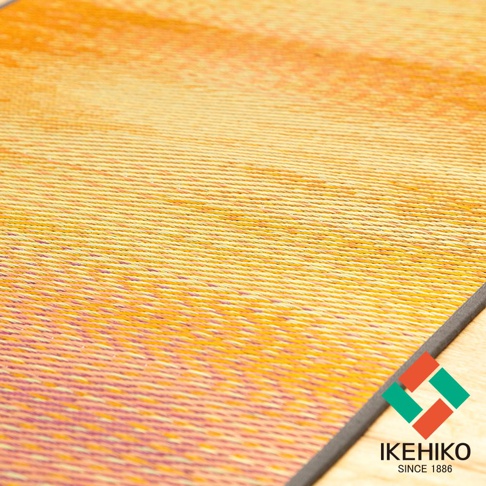 藺草編織的瑜珈墊，表面呈漸層的橘黃色與淡紫色，質地細緻，邊緣有深灰色包邊，下方露出木質地板。