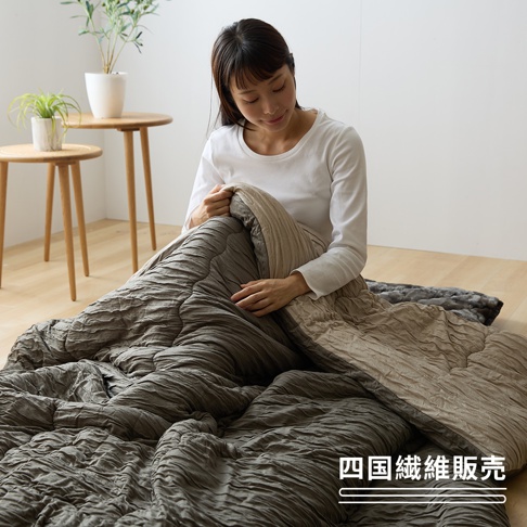 一位年輕女性坐在淺色木質地板上，手裡輕撫著一件質感柔軟、帶有天然縐褶紋理的雙色單人被，被子呈現深灰與淺棕兩種色調，背景有兩張簡約木桌與植栽盆景，圖片右下方標示有四國纖維販售的品牌字樣。