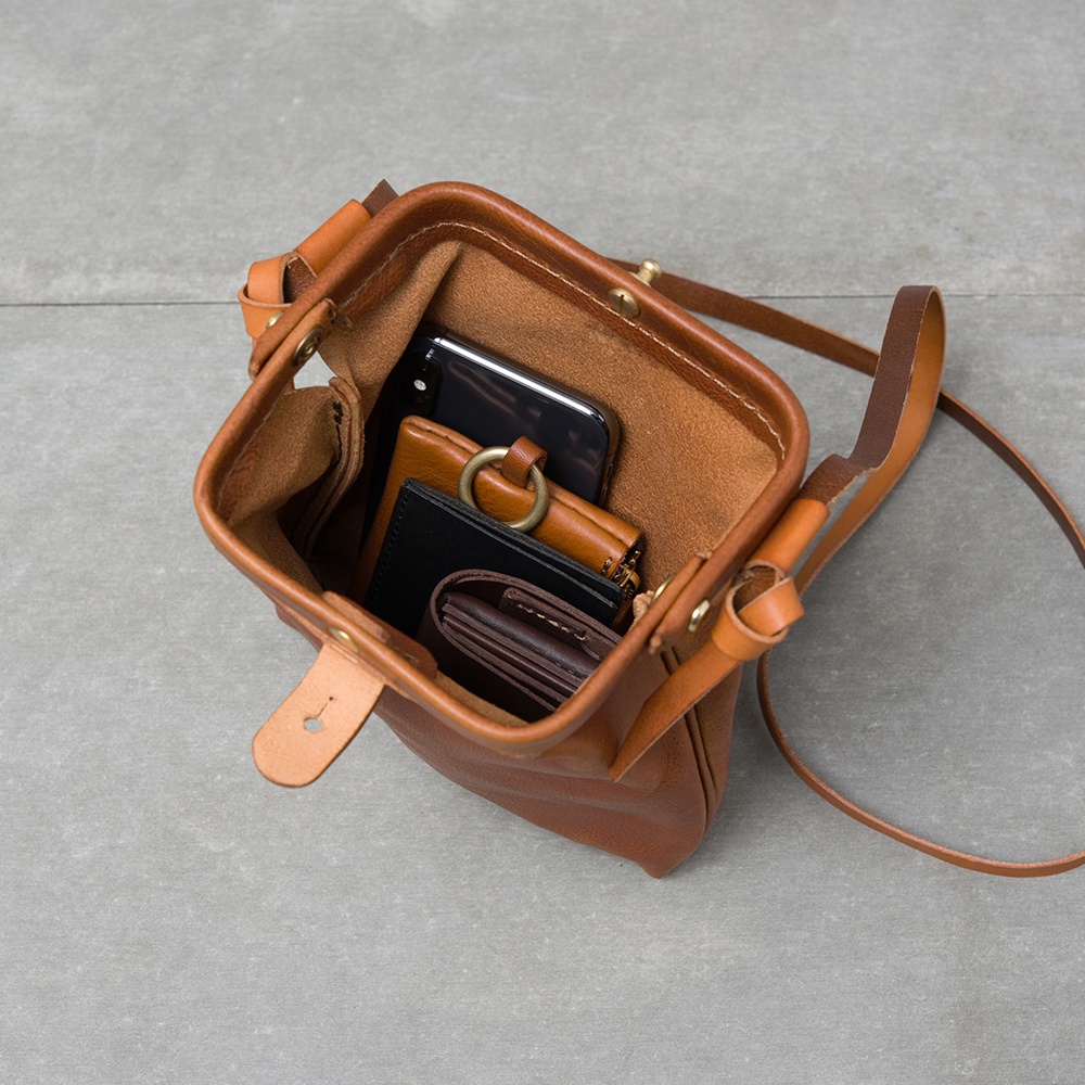 職人手作皮革Organ迷你杜勒斯側背包，採用天然棕色皮革製作，包內可見手機、短夾、卡包等物品。包身設計簡潔，配有可調節長度的肩帶。