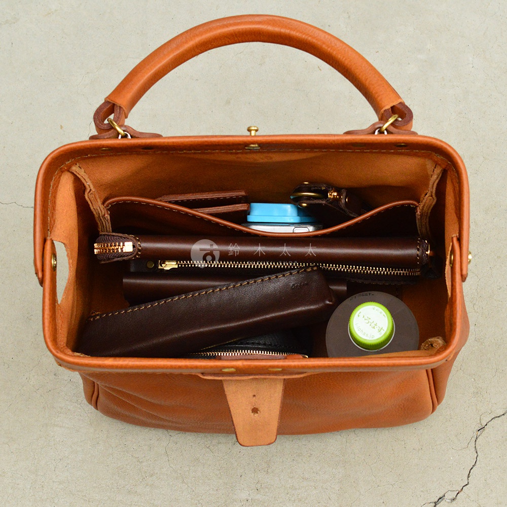 一個打開的棕褐色皮革托特包，內部裝有錢包、手機、鑰匙和一個圓形綠色罐子。