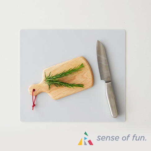 一把銀色刀具放在淺灰色砧板上，旁邊放著一個帶有紅色掛繩的木製小切菜板，上面有迷迭香。