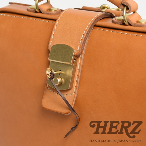 職人手作皮革杜勒斯商務後背包 BJ-10，特寫展示包款的鎖扣細節，採用天然皮革材質，復古黃銅色鎖頭，搭配淺棕色皮革與白色縫線，簡約而精緻。