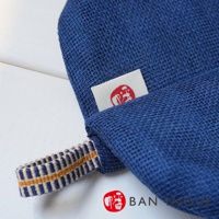 藍色蚊帳質地防燙鍋套，帶有白色織標，標籤上有紅色圖案。鍋套一側有一個藍白黃條紋編織提帶。