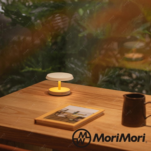 黃色底座搭配白色燈罩的LED T-Light LOUNGE工型造型燈，置於木桌上，旁邊有書本與茶壺。