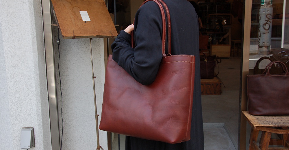 一個人背著一個大地色系的皮革托特包，包包的提把和邊緣有明顯的縫線。