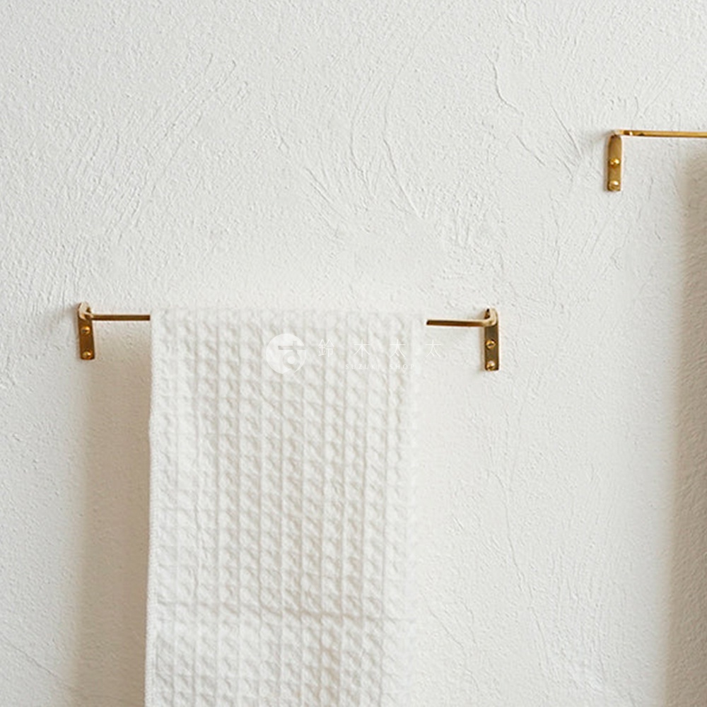 黃銅單桿毛巾架，帶有兩個黃銅支架固定在白色紋理牆壁上，一條紋格白毛巾懸掛在架子上。