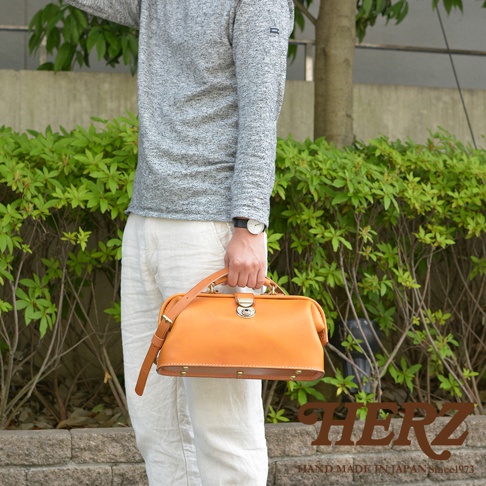 職人手作皮革長型迷你醫生包 CW-106，採用天然皮革製成，呈現溫潤的淺棕色。包款設計簡潔，配有頂部提把、可調節肩帶及金屬鎖扣，側面可見釘狀裝飾，整體散發經典復古氣息。