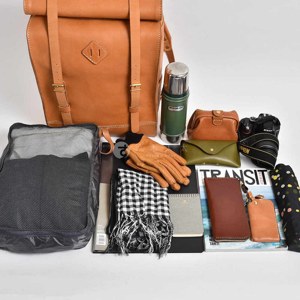 職人手作皮革露營登山用大容量後背包 R-19，搭配棕色皮革配件、綠色皮革信封包、黑色格紋圍巾、棕色皮革手套、綠色保溫瓶、黑色網狀收納袋、相機及雜誌，展現戶外冒險風格。