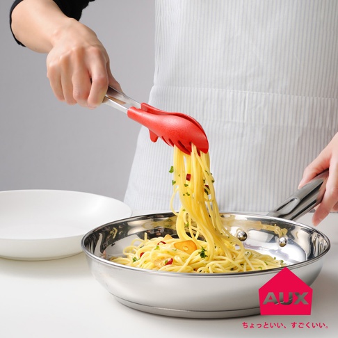 一隻紅色的矽膠料理夾夾著義大利麵，從不鏽鋼鍋中取出，鍋內有蒜片和辣椒。