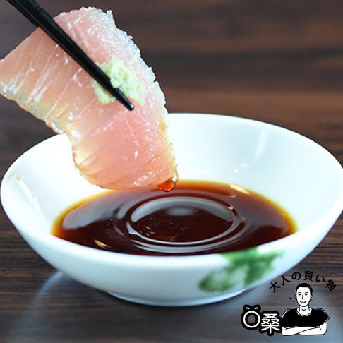 一塊生魚片被黑色筷子夾住，沾著少許綠色芥末，並正在滴入裝有深棕色醬油的白色小碟中。