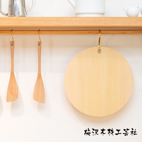 淺色木質圓形砧板，直徑約30公分，帶有一個掛孔，用金色掛鉤懸掛於木架上，旁邊是兩個木質鍋鏟。